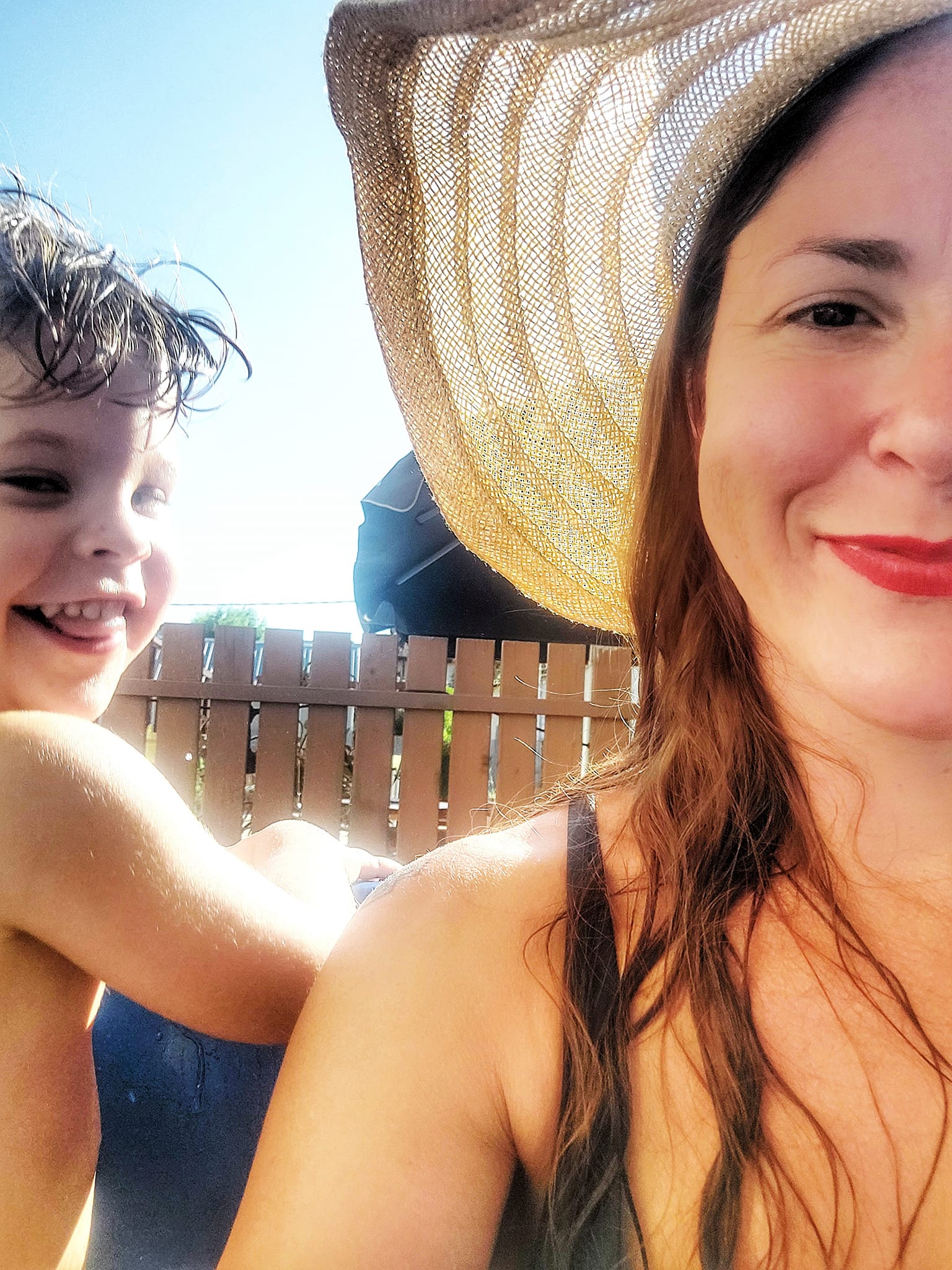 Woman in sun hat and red lipstick taking a selfie with a toddler boy looking back at the camera.