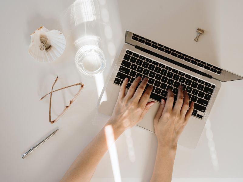 Womans hands typing on a macbook with glasses and a pen beside the computer on a white table with a stream of light coming in.